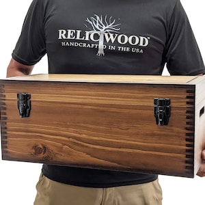 May include: A large wooden storage box with two black metal clasps. The box is made of natural wood with a dark brown stain. The box is being held by a person wearing a black t-shirt with the text "RELIC WOOD HANDCRAFTED IN THE USA".