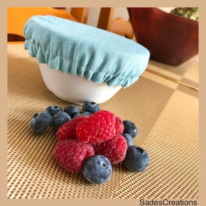 May include: A light blue reusable food cover made of fabric sits on a white bowl. The bowl is on a brown and gold patterned surface. There are blueberries and raspberries scattered around the bowl.