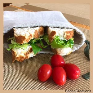 May include: Two sandwiches with lettuce and a white filling are wrapped in a beige linen cloth bag. Four red cherry tomatoes are in the foreground on a brown woven placemat.