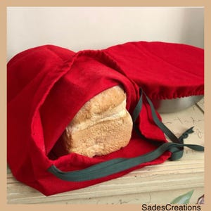 May include: Red linen bread bag with a green drawstring. A loaf of white bread is inside the bag.
