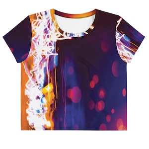 May include: A short-sleeve crop top featuring an abstract print of light trails and bokeh circles. The design displays vibrant streaks of white, orange, and yellow light against a deep purple and dark blue background, with a round neckline.