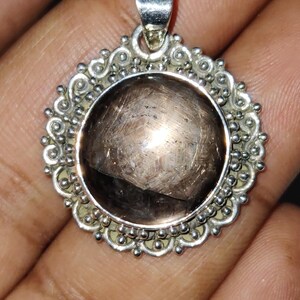 May include: A round pendant featuring a large, translucent, dark brown gemstone set in a polished silver frame. The silver setting is decorated with intricate scrollwork and small metal beads along the outer edge, topped with a silver bail.