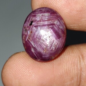 Peut inclure: Pierre précieuse ovale rouge foncé avec un motif en forme d'étoile. La surface polie reflète la lumière, révélant un motif géométrique. La pierre est tenue entre les doigts, soulignant sa taille et sa couleur.