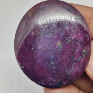 May include: A large, oval-shaped, purple gemstone with a smooth, polished surface. The stone has a swirling pattern of lighter and darker purple hues, with flecks of gold and silver throughout.