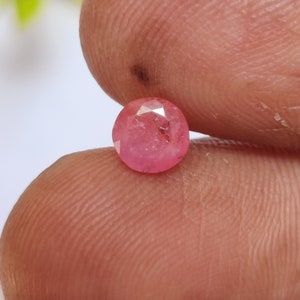 May include: A round, faceted pink gemstone held between two fingers. The gemstone has a slightly irregular shape and a light pink hue. The facets catch the light, creating a sparkling effect. The background is blurred, focusing attention on the gem.