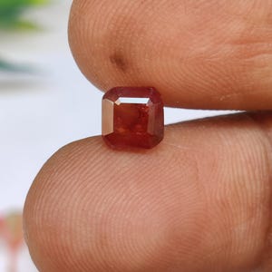 May include: A faceted, reddish-orange gemstone with an emerald cut. The gem is held between two fingers, showcasing its clarity and colour. The gem's facets reflect light, highlighting its brilliance. The overall image focuses on the gemstone's beauty and detail.