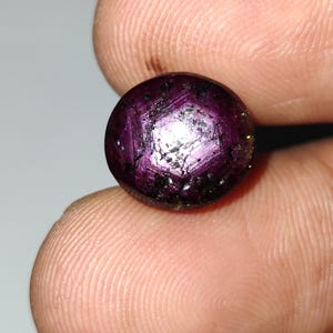 May include: A round, deep red gemstone with a star-like pattern. The gem has a polished surface with a mix of dark and light areas, and is held between two fingers. The gem is likely a ruby.