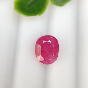 May include: A faceted, oval ruby gemstone. The ruby is a vibrant red colour with a polished surface, reflecting light. The gemstone is set against a white background, with green leaves in the upper portion of the image.