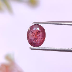 May include: An oval-shaped, faceted ruby gemstone held by silver tweezers. The ruby has a deep red color with darker inclusions, and is set against a soft, blurred background. The gemstone is a focal point, showcasing its natural beauty and potential for jewelry.