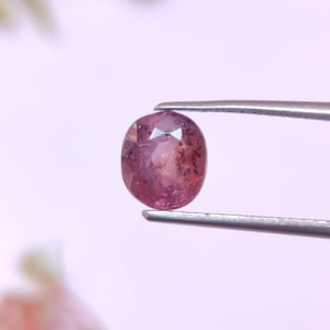May include: A faceted, oval-shaped, pink gemstone is held by a pair of silver tweezers. The gem has a mix of pink and brown hues, with visible internal inclusions. The background is a soft, blurred white.