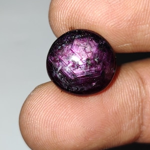 May include: A round, polished gemstone with a deep purple hue, exhibiting a star-like effect. The gem has a smooth, reflective surface with internal crystalline structures. The stone is held between fingers, showcasing its size and color.