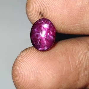May include: An oval-shaped, deep magenta-colored gemstone with a cabochon cut. The stone displays a distinct six-rayed star effect, known as asterism, across its polished surface, with visible internal inclusions and a translucent, glossy texture.