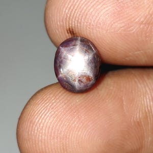 May include: Oval-shaped, star sapphire gemstone. The gem displays a purplish-gray hue with a prominent six-ray star effect. The stone's surface reflects light, creating a shimmering appearance. The gemstone is held between two fingers.