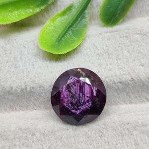 Könnte beinhalten: Ein runder, facettierter Edelstein in tiefem Violett und Magenta, der innere kristalline Strukturen und lichtreflektierende Oberflächen zeigt. Der Stein hat eine polierte Oberfläche mit mehreren dreieckigen Facetten.