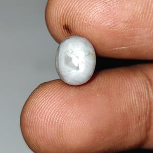 May include: A small, oval-shaped, white gemstone with a pearlescent sheen. The gem is held between two fingers, showcasing its smooth, polished surface. The stone's color is primarily white with subtle gray and cream tones.