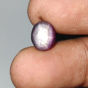 Pode incluir: Uma pedra preciosa oval polida, com um tom rosa-púrpura. A gema tem um efeito de estrela em sua superfície, com a luz refletindo nas facetas. A pedra é segurada entre dois dedos, mostrando seu tamanho e cor.