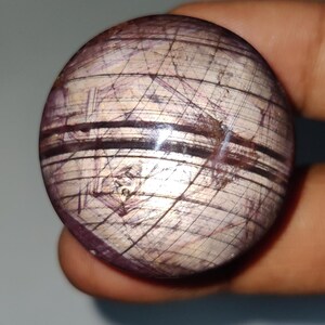 Puede incluir: Una piedra esférica pulida que presenta una base de color beige claro con prominentes inclusiones lineales de color púrpura oscuro y negro que crean un patrón de cuadrícula en la superficie. La esfera tiene un acabado liso y reflectante con texturas naturales visibles.