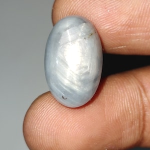 May include: A pale blue, oval gemstone with a star effect. The polished surface reflects light, creating a shimmering appearance. The gem is held between fingers, showcasing its size and shape.