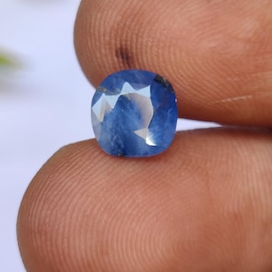 May include: A faceted, oval-shaped sapphire gemstone. The sapphire is a deep, vibrant blue color with internal reflections. The gemstone is held between two fingers, showcasing its clarity and brilliance. The gem is a natural, untreated stone.