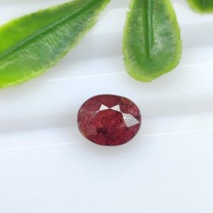 Può includere: Una pietra preziosa ovale sfaccettata di colore rosso intenso con inclusioni interne visibili. La pietra presenta una superficie lucida con molteplici sfaccettature geometriche che riflettono la luce, mostrando una tonalità cremisi scuro.