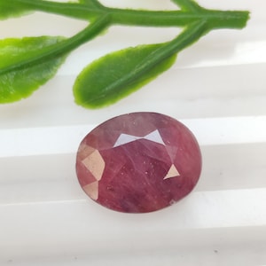 Pode incluir: Gema de rubi facetada em forma oval. A gema é de cor rosa-avermelhada profunda com múltiplas facetas que refletem a luz. Um ramo verde com folhas está no fundo.