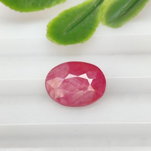 May include: A faceted, oval-shaped ruby gemstone. The ruby is a deep, vibrant red color with multiple facets that reflect light. The gemstone is set against a white background, with green leaves in the background.