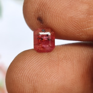 Puede incluir: Una piedra preciosa de corte rectangular facetado con un tono rosa rojizo vibrante. La piedra presenta un cuerpo transparente con inclusiones oscuras y dispersas visibles, una superficie pulida y bordes geométricos definidos.