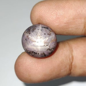 Puede incluir: Una piedra preciosa de zafiro estrella redonda y púrpura con una estrella de seis puntas en el centro. La piedra preciosa está tallada en cabujón y tiene una superficie lisa y pulida.