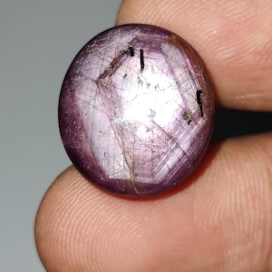 May include: A circular, polished gemstone in a deep purplish-red colour. The stone has a smooth, reflective surface with internal fractures and inclusions. The gemstone is held between two fingers, showing its size and colour. The gem appears to be a ruby.