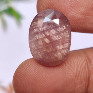 May include: Oval-shaped, faceted gemstone with a pale pink hue. The gem has internal inclusions and a polished surface, reflecting light. The stone is held between fingers, showcasing its size and shape. Ideal for jewelry making.