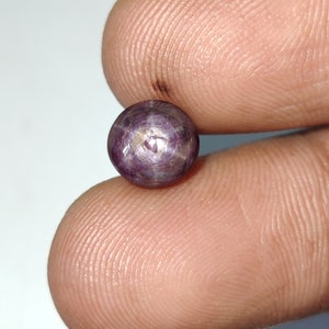 Puede incluir: Una piedra preciosa redonda de color púrpura con un patrón en forma de estrella en el centro. La piedra preciosa mide aproximadamente 1,25 cm de diámetro.