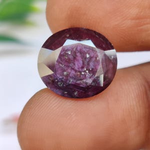 Puede incluir: Una gema facetada, de corte ovalado, de color púrpura intenso. La gema tiene una superficie reflectante con inclusiones internas, sostenida entre dos dedos. Las facetas de la gema capturan la luz, creando un efecto brillante.
