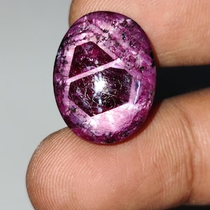 Könnte beinhalten: Ein ovaler Cabochon-Edelstein in tiefen Magenta- und Violetttönen mit einem markanten, dunklen, geometrischen hexagonalen Einschluss in der Mitte. Die Oberfläche weist eine polierte, reflektierende Textur mit natürlichen inneren Adern auf.