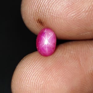 Op de afbeelding: Een levendige, ovaalvormige, roze edelsteen met een stervormig patroon. De edelsteen heeft een diepe magenta kleur en lijkt gepolijst. Het ster-effect wordt gecreëerd door licht dat weerkaatst op insluitsels in de steen. Dit is waarschijnlijk een robijn.