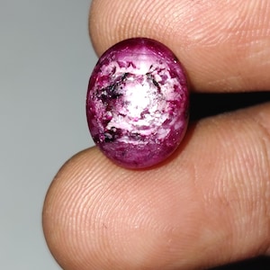 May include: An oval-shaped gemstone, polished to a high sheen, displaying a rich magenta colour with internal white and black markings. The gem is held between fingers, revealing its size and texture. The surface reflects light, accentuating its unique patterns.
