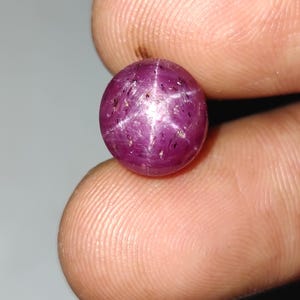 May include: A round, polished ruby gemstone with a star effect. The gem is a deep purplish-red color, with a six-rayed star pattern visible across its surface. The ruby is held between two fingers, showcasing its size and shape.