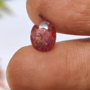 Puede incluir: Una gema facetada de forma ovalada con un tono rosa rojizo. La gema tiene inclusiones internas y una superficie pulida, sostenida entre los dedos. El color de la piedra varía de rosa claro a oscuro, con algunas áreas translúcidas.