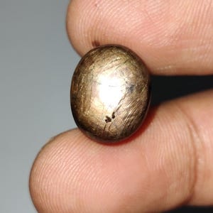 Puede incluir: Una gema ovalada pulida, de color marrón oscuro, con un patrón reflectante en forma de estrella. La piedra se sostiene entre dos dedos, mostrando su tamaño y textura. La superficie presenta inclusiones y marcas naturales.