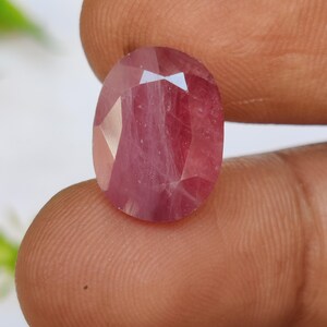 Könnte beinhalten: Ein oval geschliffener Rubin-Edelstein. Der Rubin hat eine tiefrosa-rote Farbe mit sichtbaren inneren Einschlüssen. Der Edelstein wird zwischen den Fingern gehalten, wodurch seine polierten Oberflächen und die Lichtbrechung hervorgehoben werden.