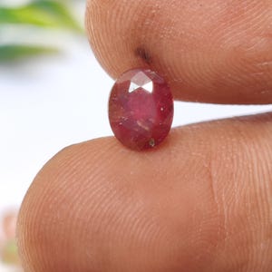 Puede incluir: Una gema de rubí facetada, de forma ovalada. El rubí es de color rojo intenso con inclusiones visibles. Se sostiene entre dos dedos, mostrando su tamaño y forma. El fondo está borroso, resaltando la gema.