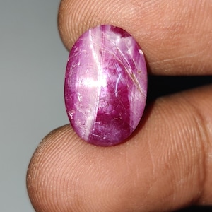 May include: An oval-shaped, cabochon-cut star ruby gemstone with a deep magenta-red color. The stone features a distinct, light-colored six-rayed star effect across its surface, along with visible internal inclusions and natural fractures.