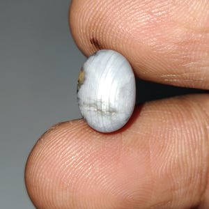 May include: A small, oval-shaped gemstone with a pearly white color and subtle gray and yellow inclusions. The polished surface reflects light, showcasing a natural, layered pattern. Held between fingers, the gem appears to be a cabochon, suitable for jewelry.