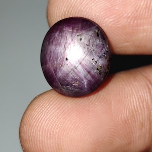 Può includere: Una gemma ovale lucidata di colore viola intenso. La pietra presenta un effetto stella, con la luce che si riflette sulla sua superficie. Piccole inclusioni scure sono visibili all'interno della gemma, aggiungendo al suo carattere unico.
