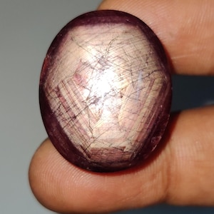Pode incluir: Uma pedra preciosa em formato de cabochão oval com um tom vermelho-púrpura profundo. A superfície exibe um efeito de estrela de seis raios, conhecido como asterismo, com luz refletida sobre a cúpula polida e translúcida.
