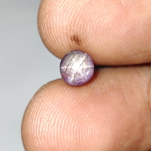 Peut inclure: Une petite pierre précieuse ronde, rose-violet, avec un motif en forme d'étoile de lignes entrecroisées sur sa surface. La gemme est tenue entre deux doigts, mettant en valeur sa taille et ses détails. La surface polie de la pierre reflète la lumière.