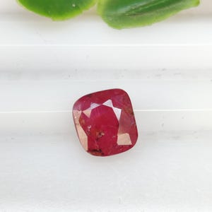 May include: A faceted, cushion-cut ruby gemstone. The gem is a deep red color with internal inclusions and a polished surface. The background is white with a green leaf in the upper corner.