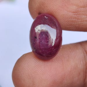 May include: A polished, oval-shaped ruby gemstone. The gem is a deep, rich red color with a smooth, reflective surface. The ruby is held between two fingers, showcasing its size and shape.