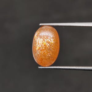 May include: An oval-shaped, orange sunstone gemstone held by tweezers. The gemstone has a sparkling, glittery appearance with flecks of gold and orange throughout. The background is a solid, dark gray.