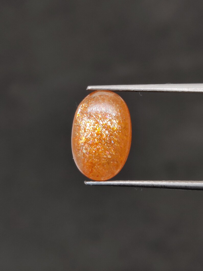 May include: An oval-shaped, orange sunstone gemstone held by tweezers. The gemstone has a sparkling, glittery appearance with flecks of gold and orange throughout. The background is a solid, dark gray.