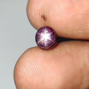 May include: A small, round, purplish-red gemstone with a star-like pattern. The gem is held between two fingers, showcasing its polished surface and unique optical effect. The gemstone appears to be a ruby.